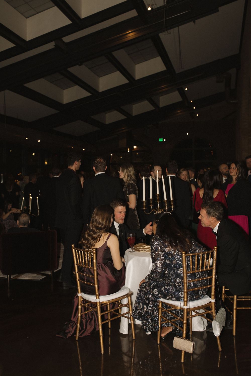 Guests mingle at an elegant evening event with tall candlesticks and formal table settings in a dimly lit venue.