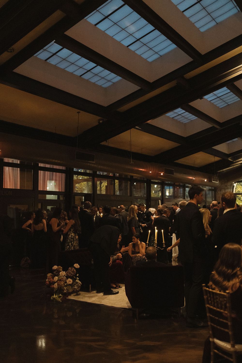 Dimly lit indoor event space with skylights and guests mingling at tables during an evening gathering.