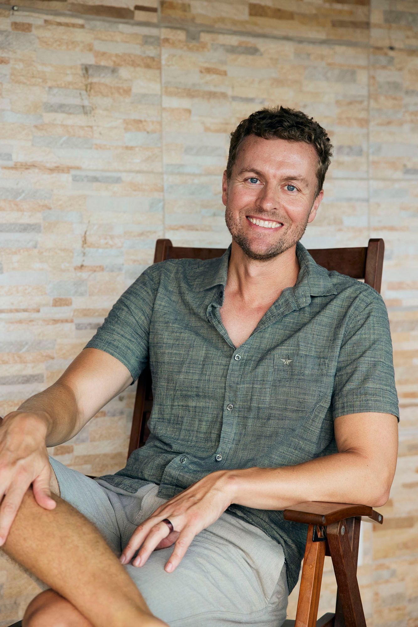 A person wearing a gray polo shirt sits casually in a wooden chair against a textured stone wall.