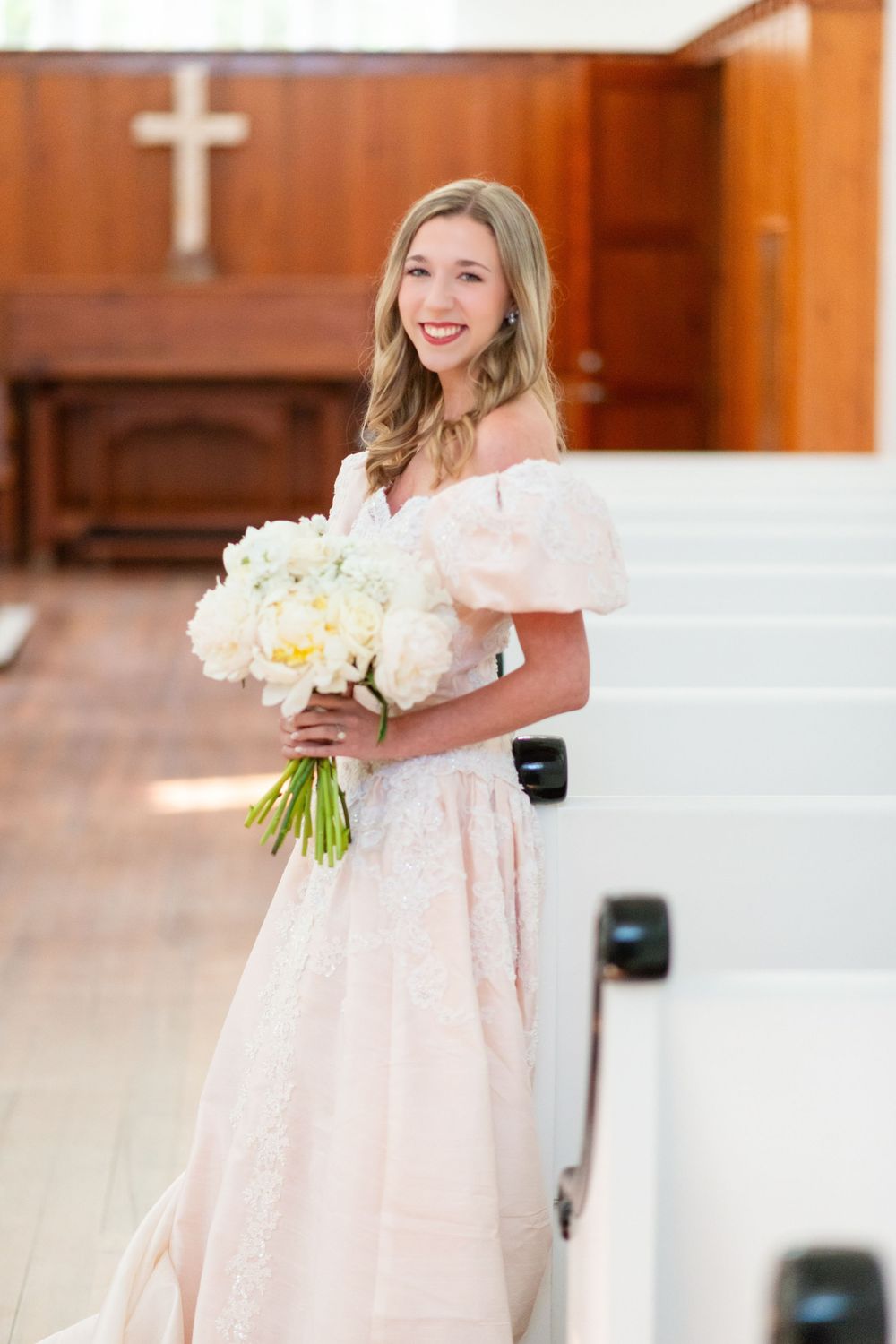 A bride poses with white bouquet in an off-shoulder gown inside a church with wooden paneling.