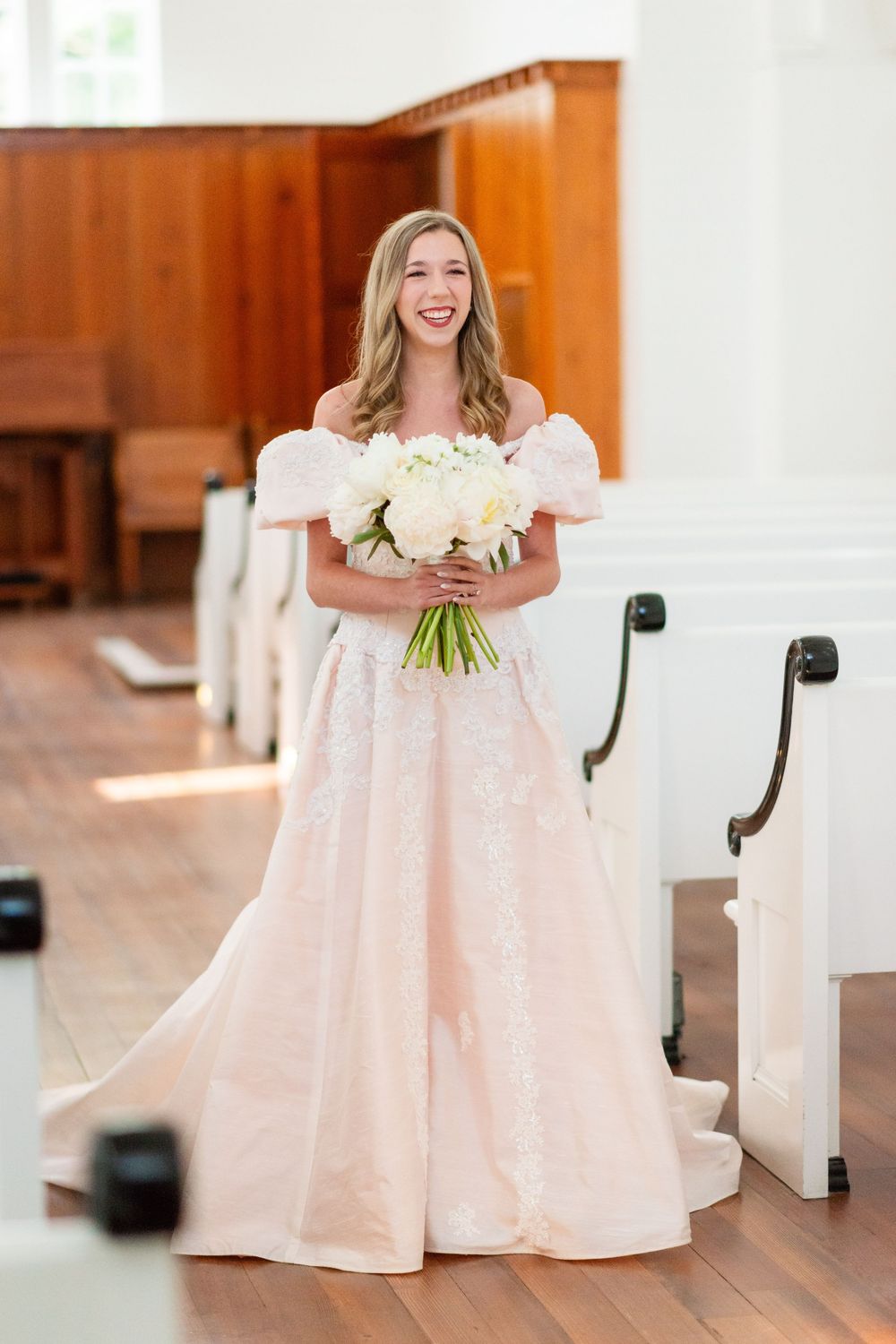 Person in a blush pink formal gown holding white flowers in a church interior.