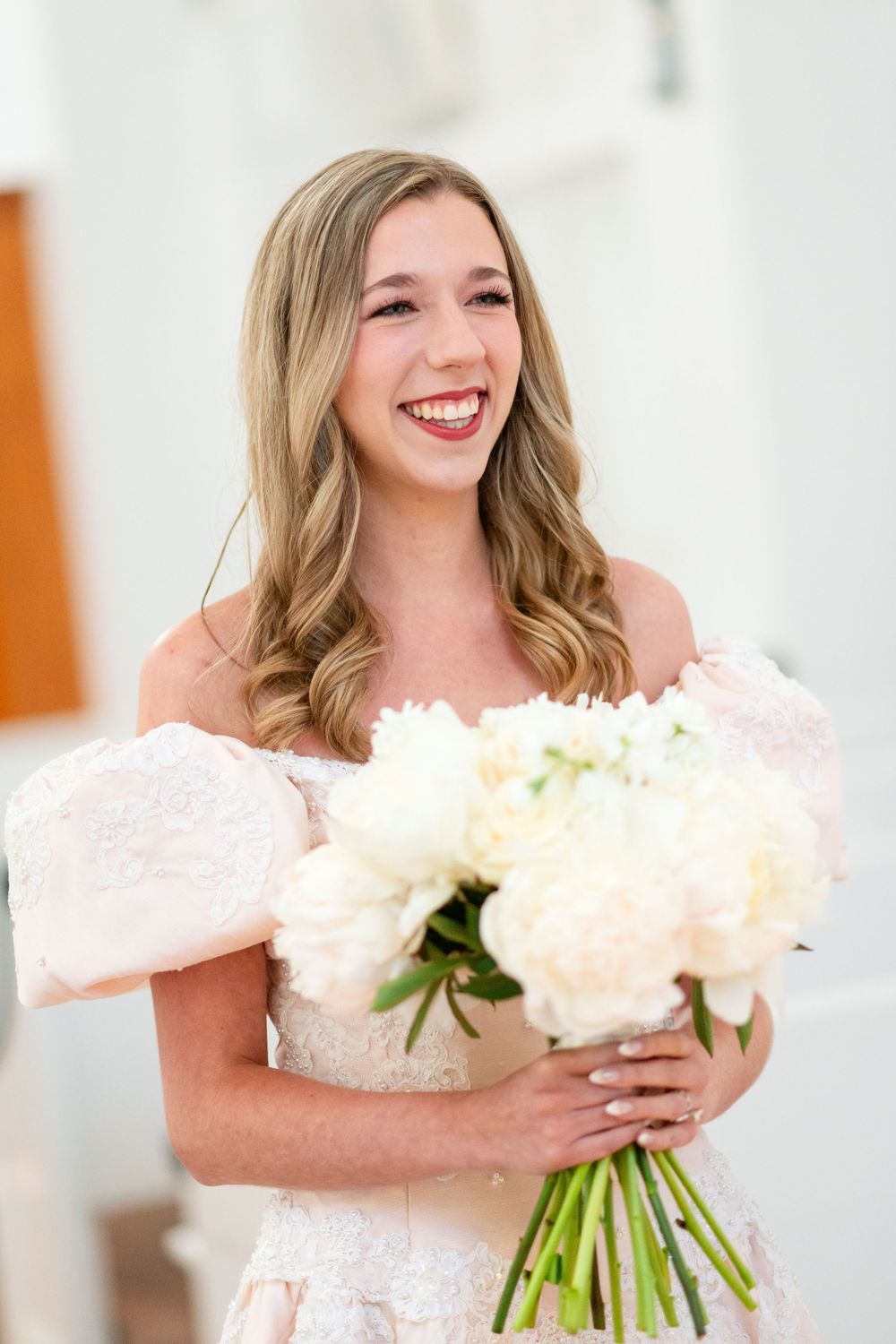 White wedding bouquet held in front of a bright white chapel interior.
