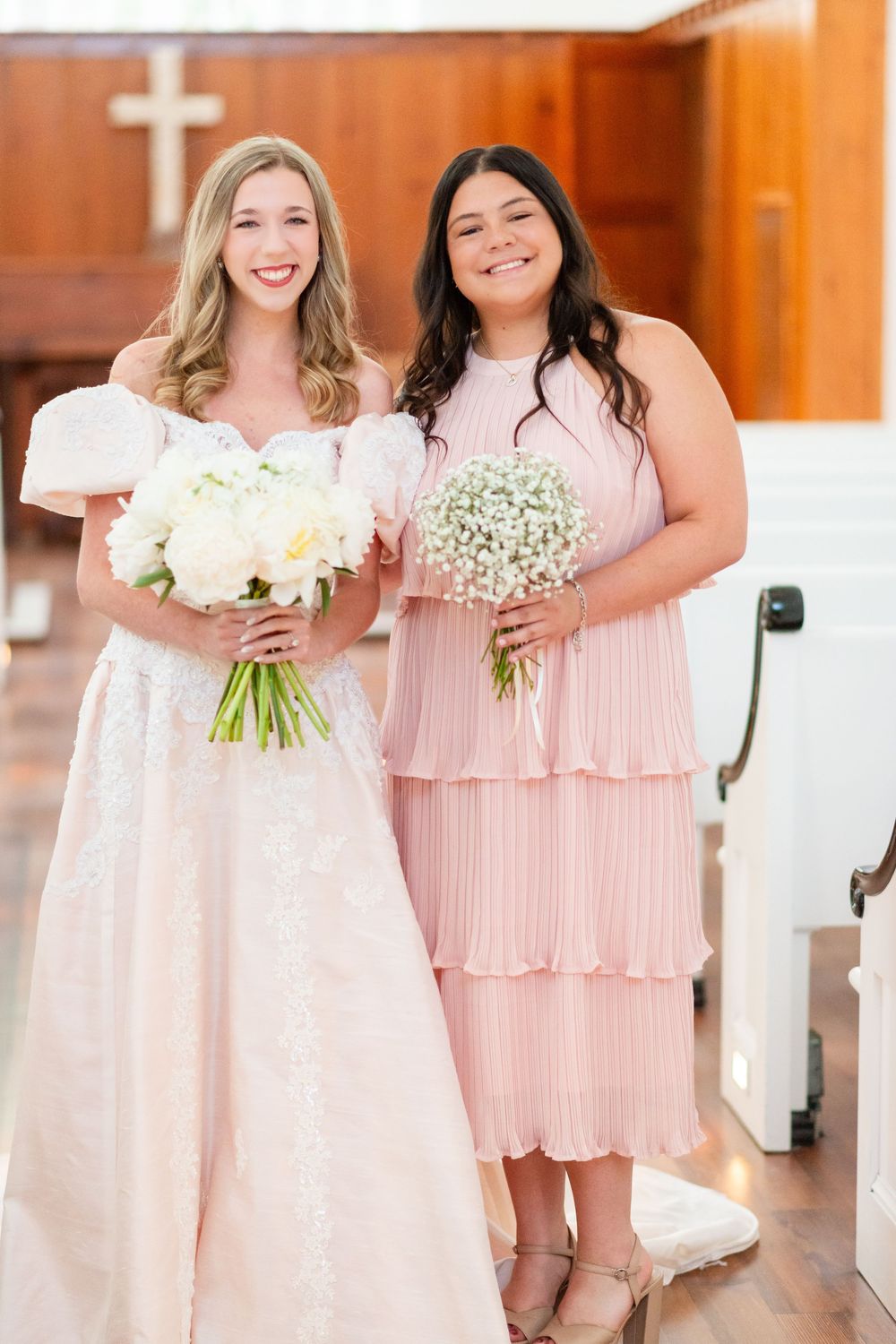 Two wedding attendants pose in a church holding white and baby's breath bouquets.