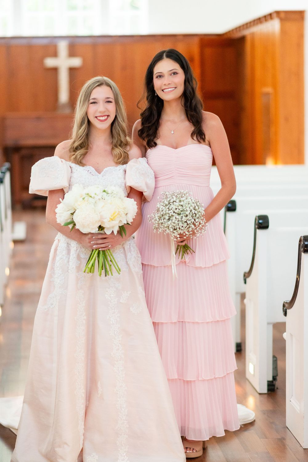 Bridesmaids in light pink dresses holding white bouquets stand together in a church setting.