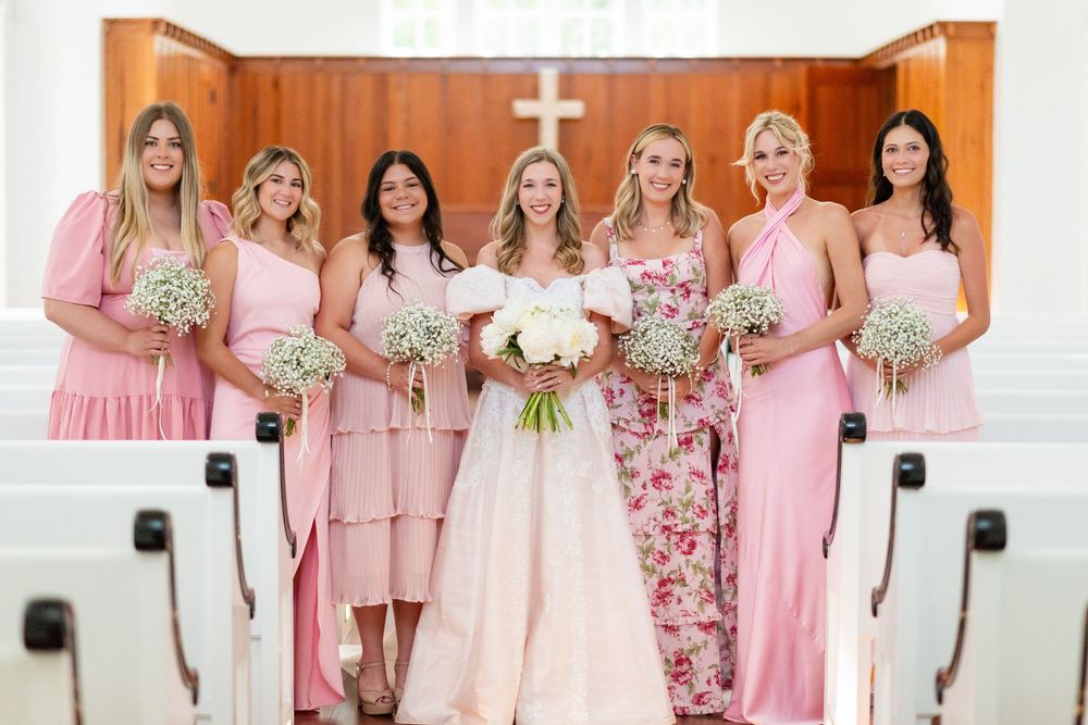 Bridesmaids in various pink dresses pose together in a church with their bouquets.