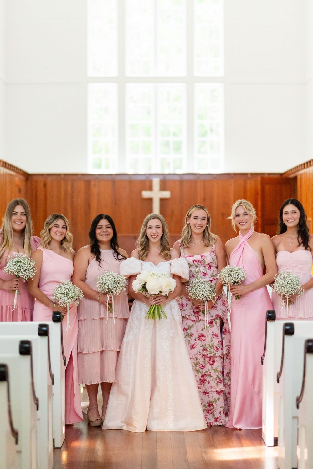 Bridesmaids in pink dresses holding baby's breath bouquets pose in a church with wooden panels.