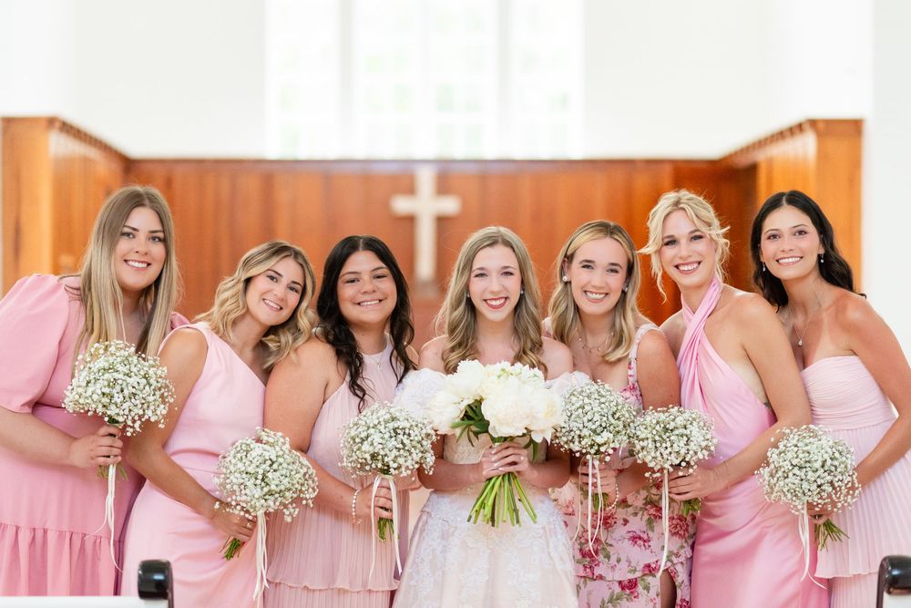 Bridesmaids in pink dresses hold baby's breath bouquets while standing in front of a white church.