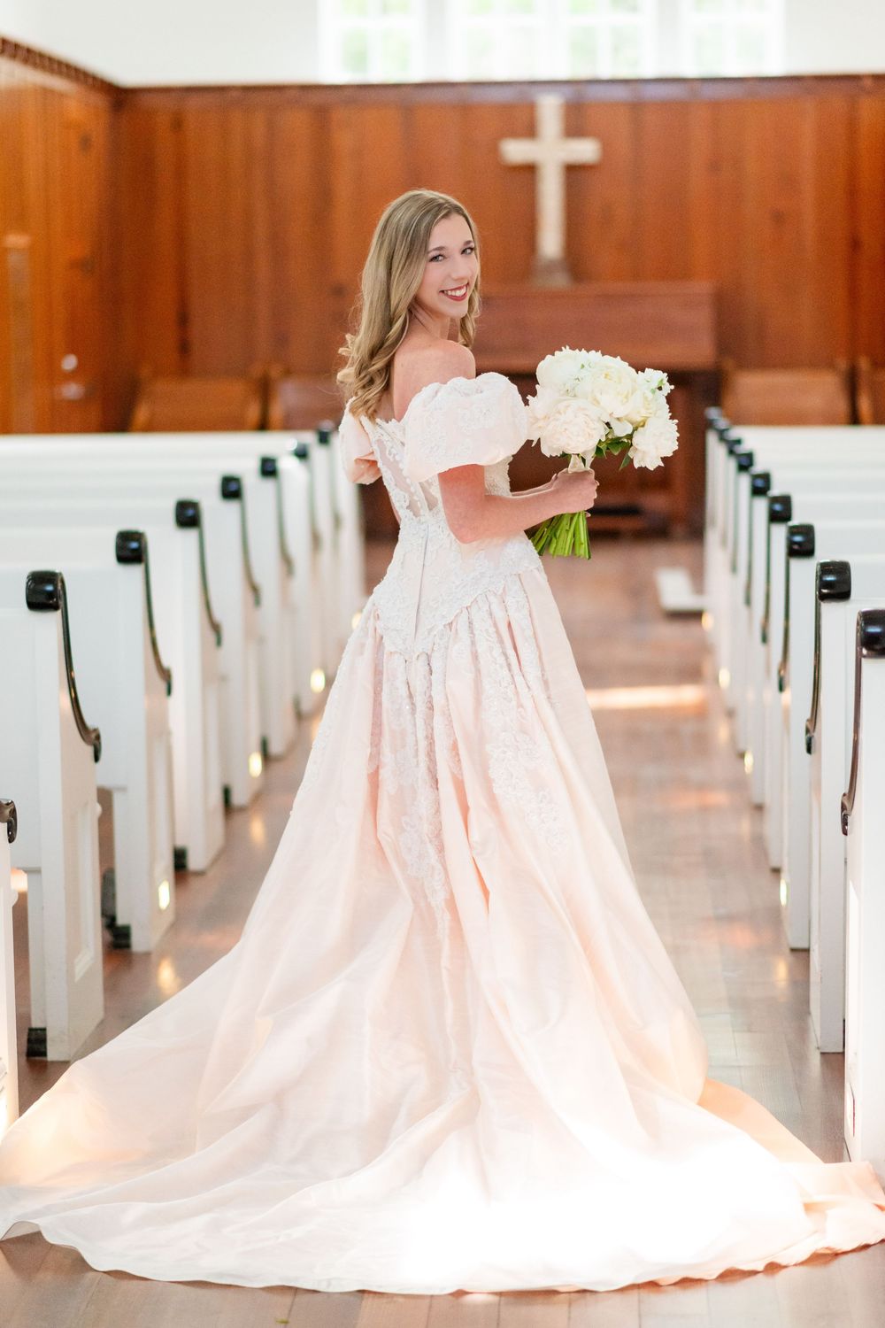 A bride in an off-shoulder blush wedding gown holds white flowers in a traditional church setting.
