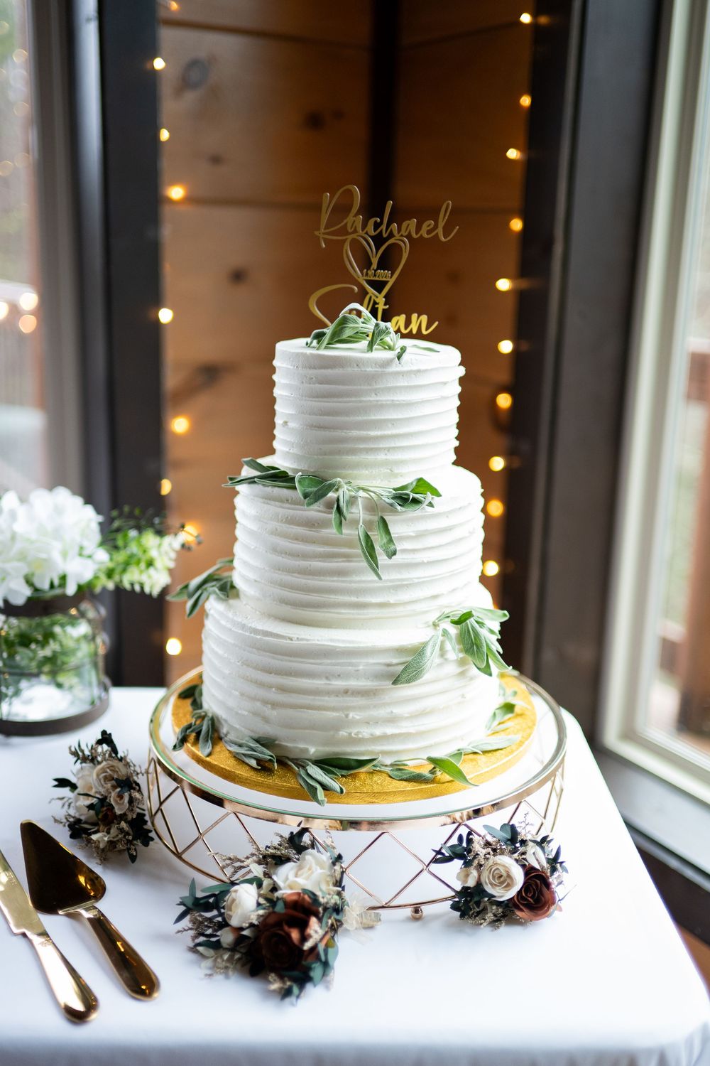 Three elegant white wedding cakes decorated with greenery and flowers displayed on gold stands against a lit backdrop.
