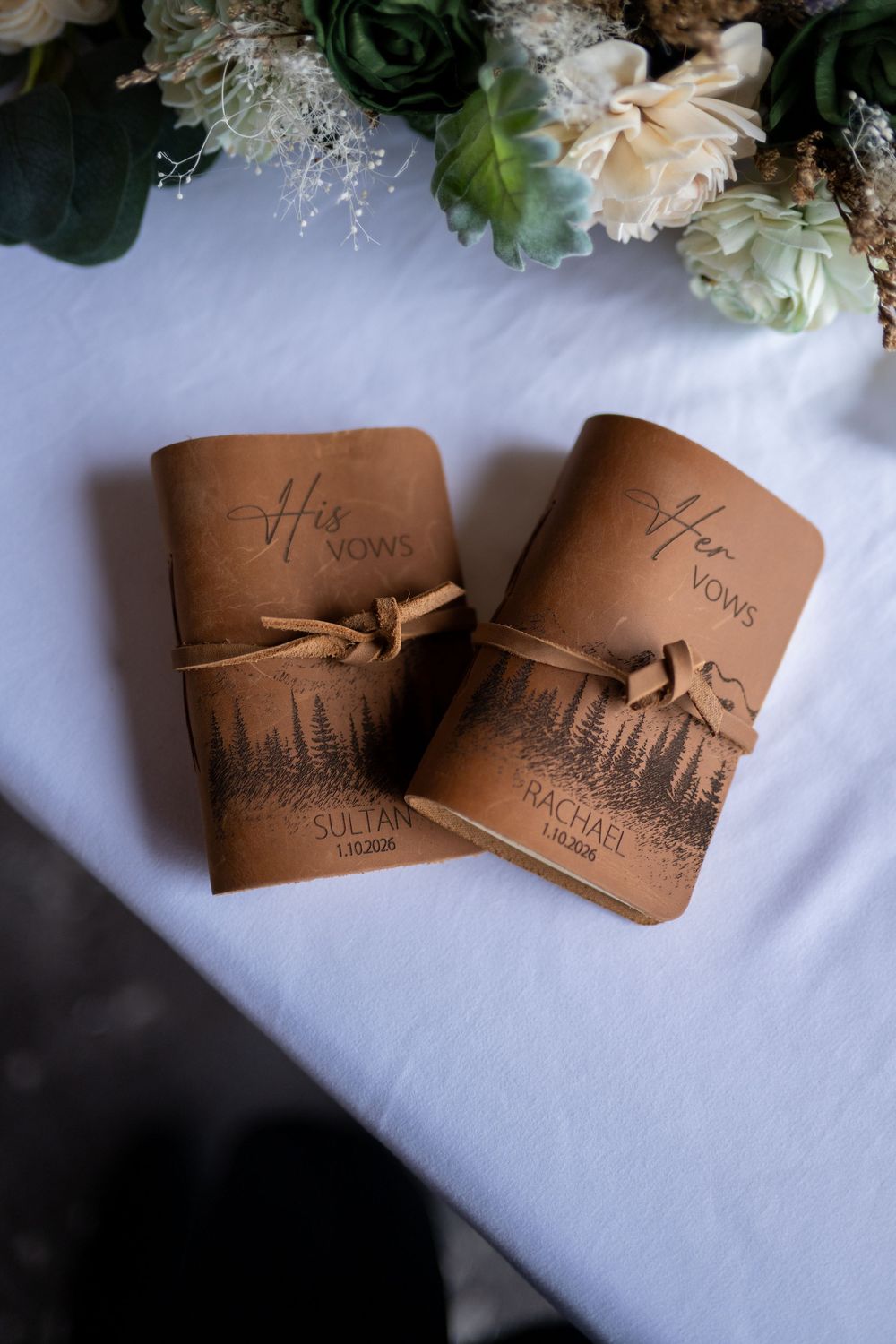 Brown leather wedding vows books with cityscape design and bow details displayed on white surface with floral accents.