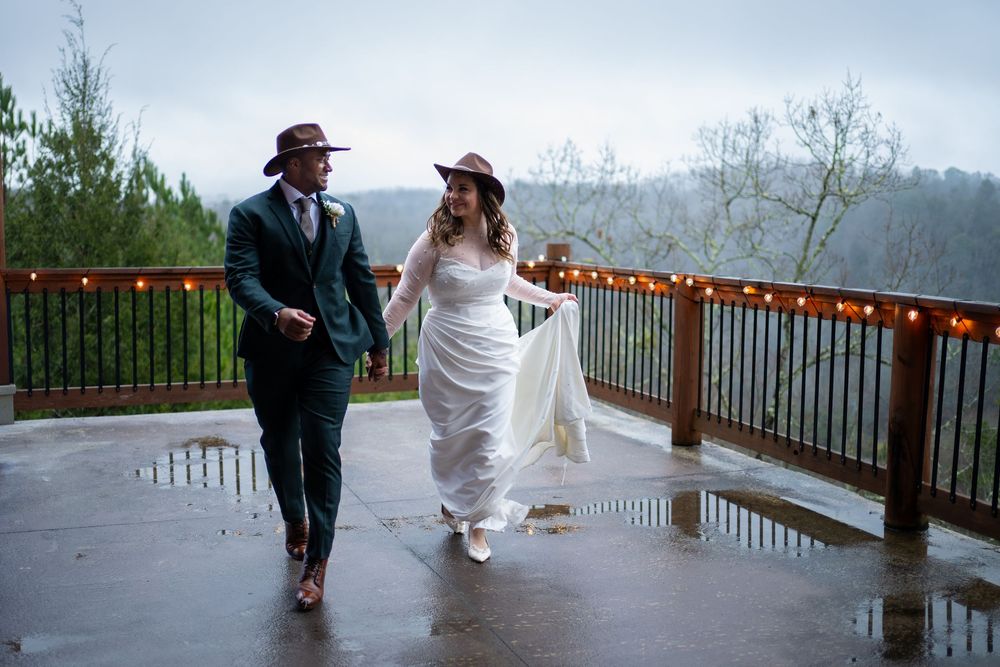 A couple in wedding attire walks together along a rustic wooden deck overlooking a scenic mountain landscape.