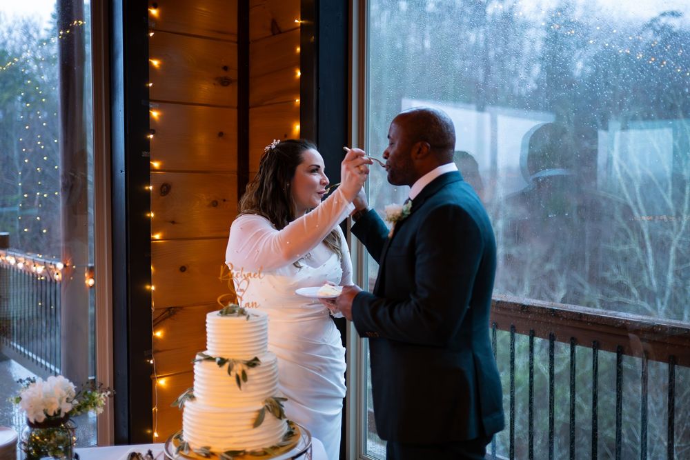 A couple shares their first bite of wedding cake together on a porch decorated with string lights and scenic views.