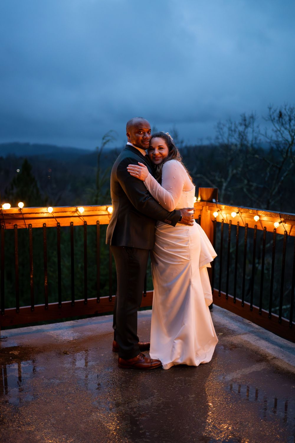 A romantic couple embraces on a rainy evening balcony with glowing string lights creating a magical atmosphere.