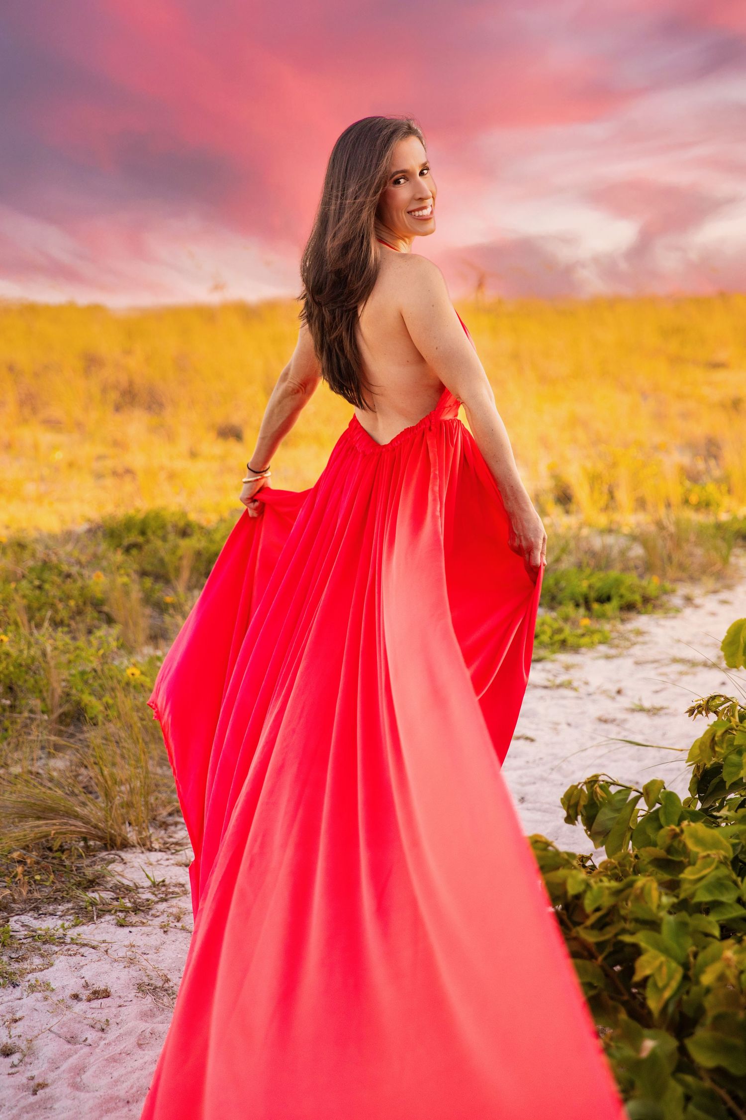 A flowing red dress catches the wind against a dramatic sunset sky on a sandy beach.