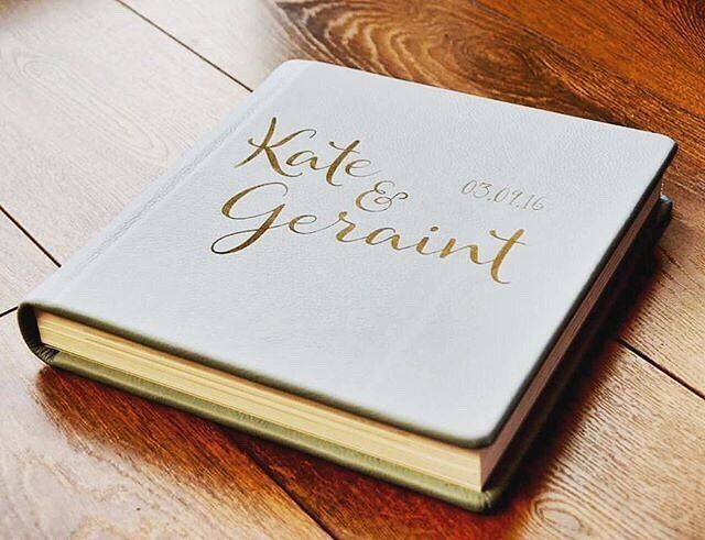 Wedding album with gold calligraphy text on white leather cover placed on wooden surface.