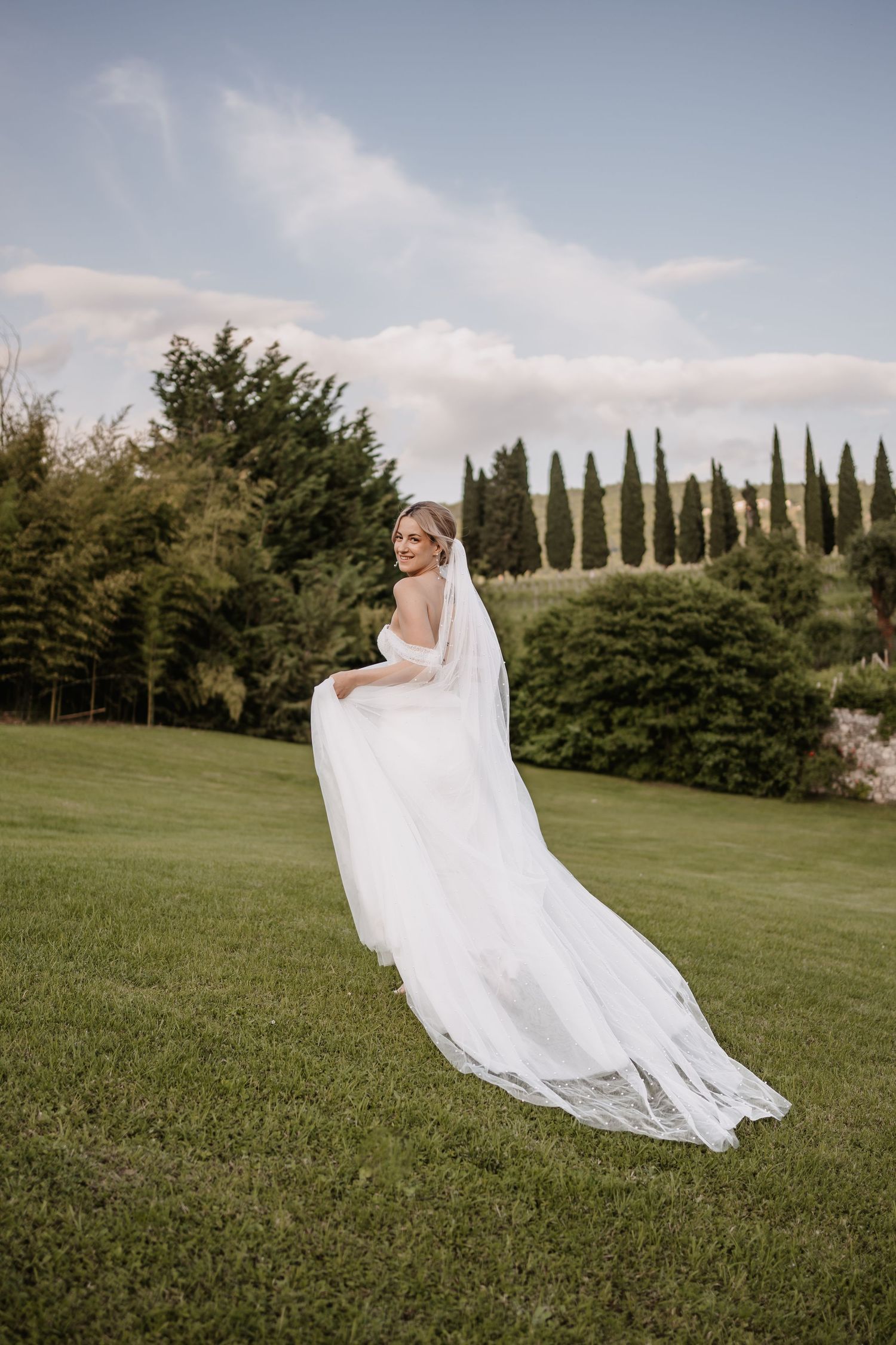 A bride in a flowing white dress with a large bow looks back while walking on a garden path with her partner.