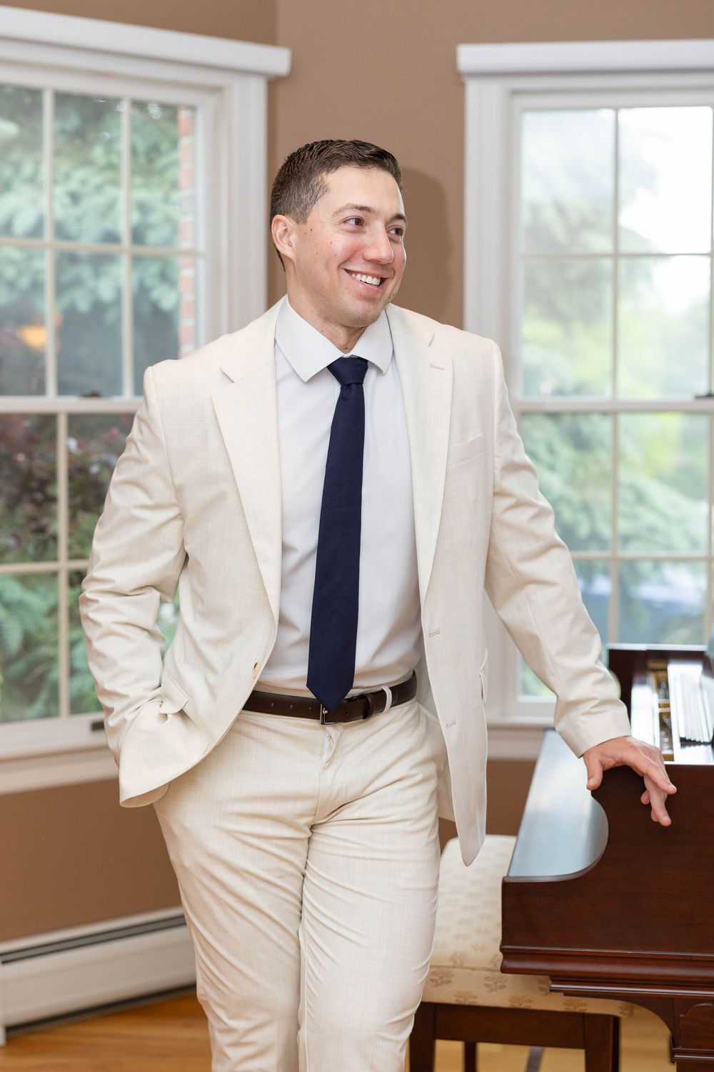 A person in a cream-colored suit and navy tie stands casually in a room with large windows.