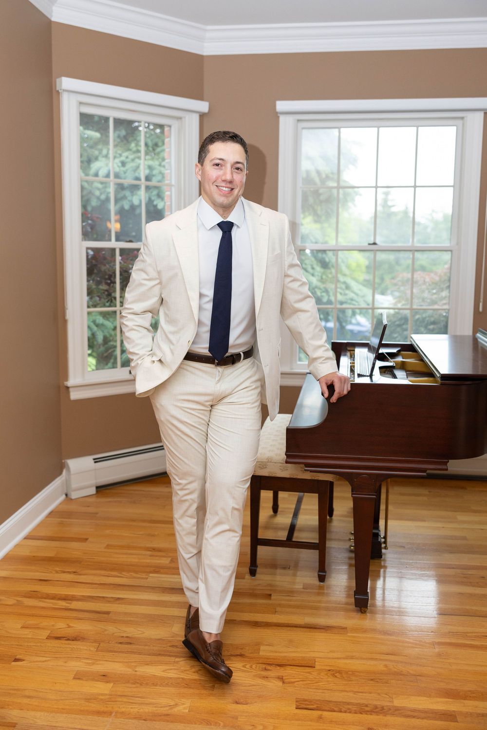 A person in a cream-colored suit and blue tie poses playfully in a bright room with hardwood floors and a grand piano.