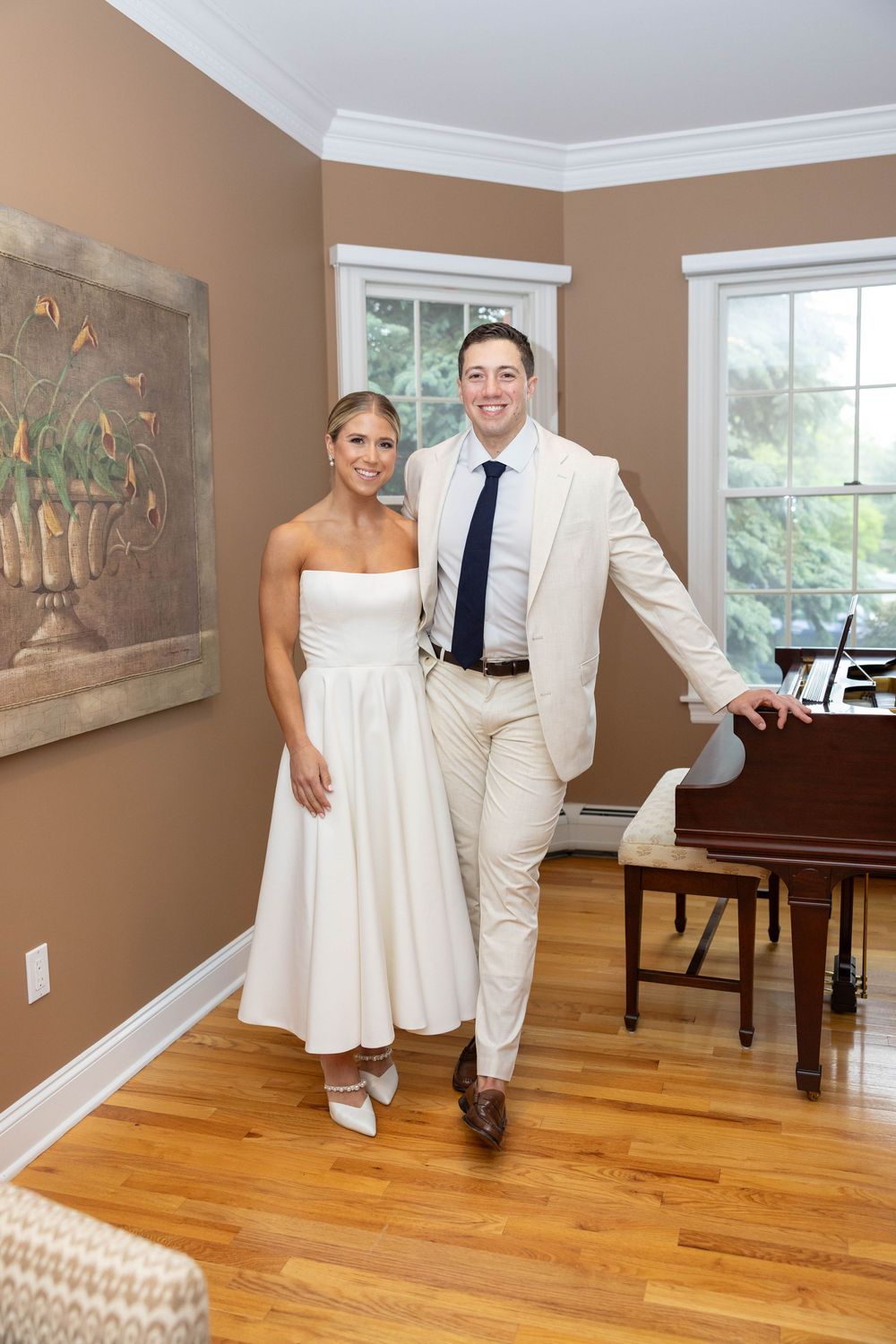 A couple in elegant white attire pose together in a room with hardwood floors and a piano.