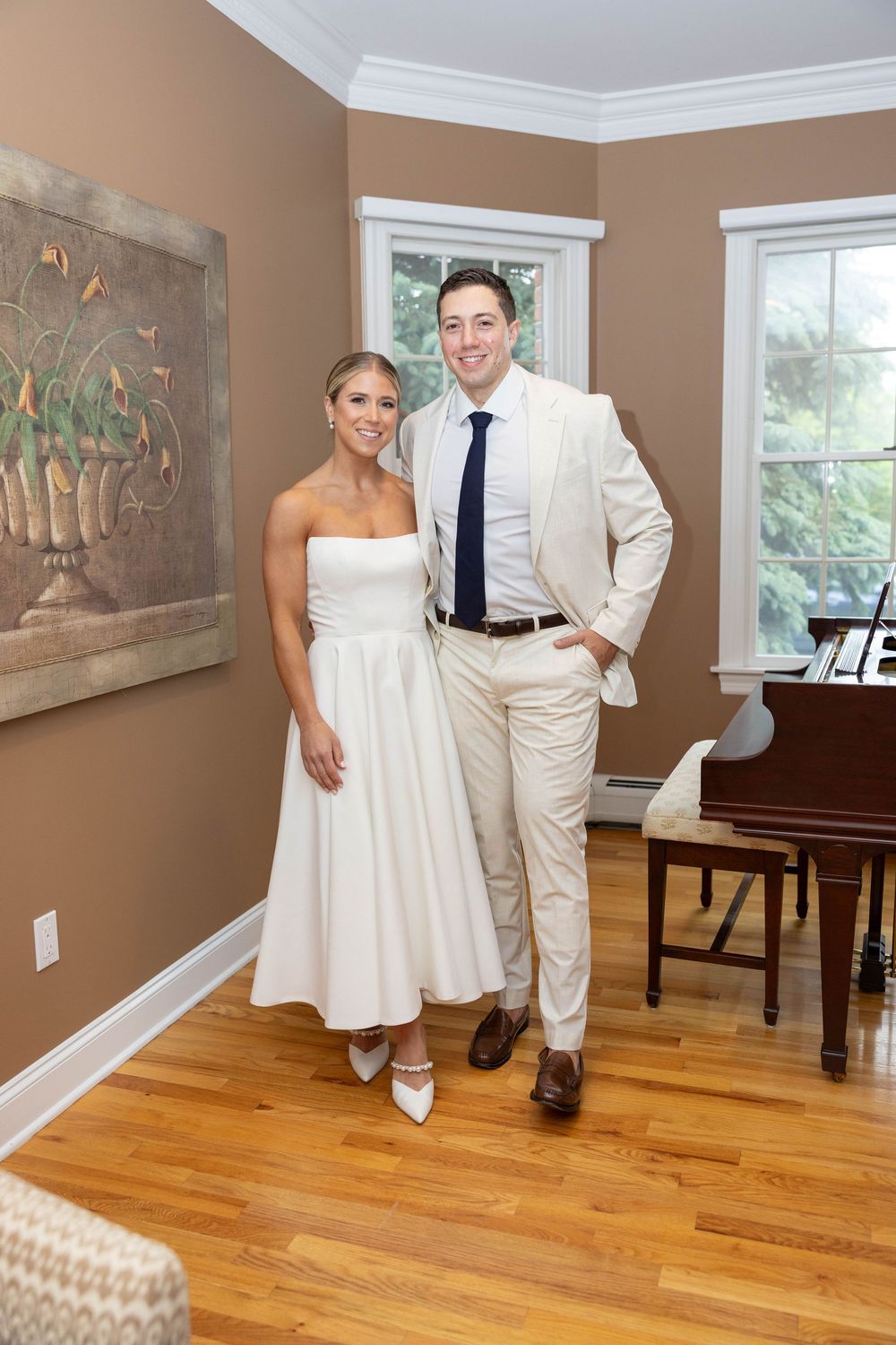 A couple in wedding attire stands together in a room with brown walls and hardwood floors near a piano.