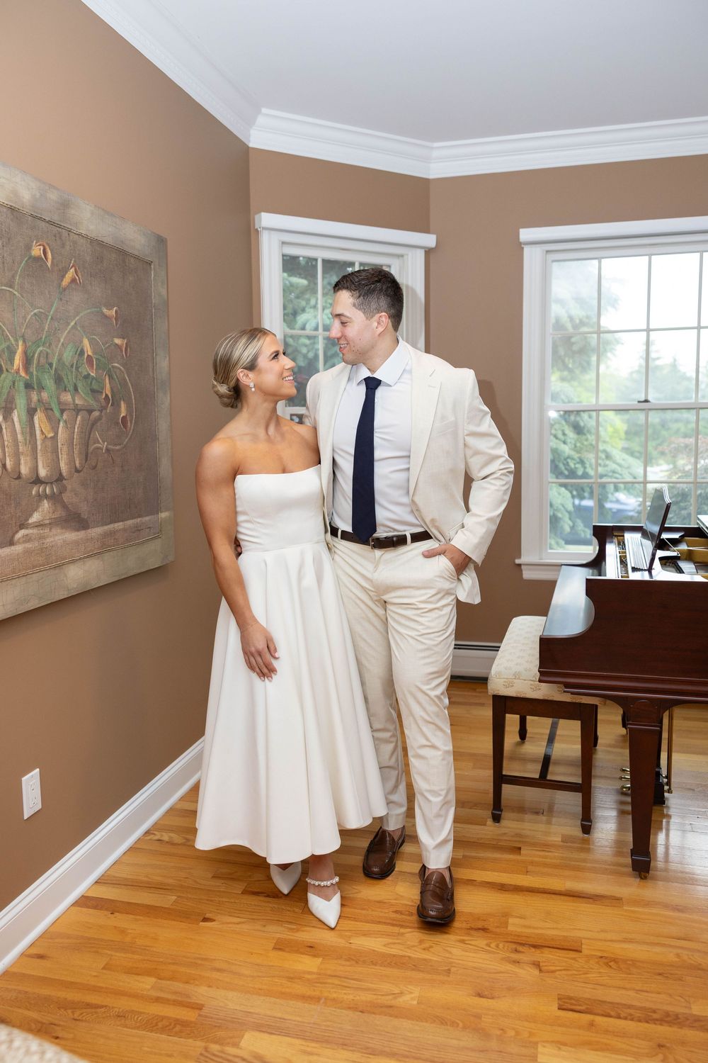 A couple in white formal attire share intimate moments in an elegant room with hardwood floors and artwork.