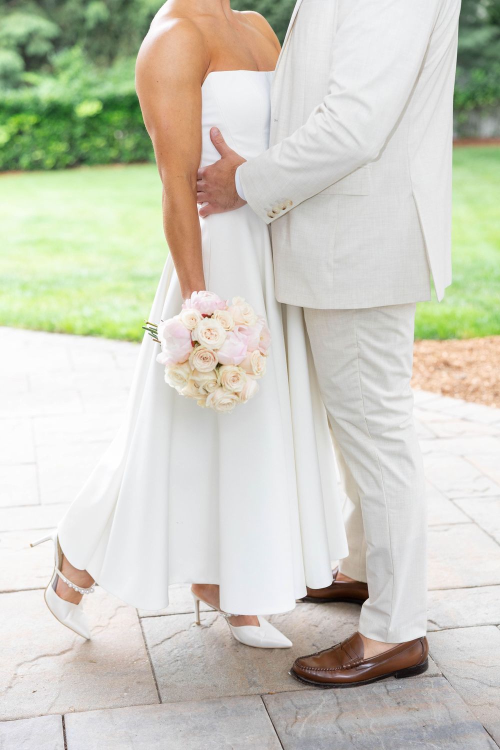A couple in wedding attire standing close together on stone pavers, with the bride holding a pink and white bouquet.