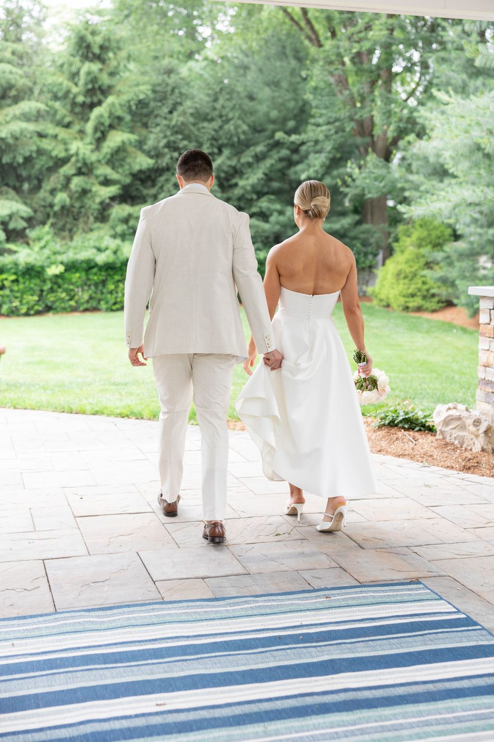A couple in wedding attire walking away on a stone patio with blue striped steps in a garden setting.