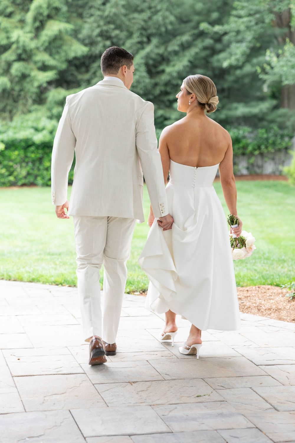 A bride and groom walk away from camera on stone patio, surrounded by lush greenery on their wedding day.