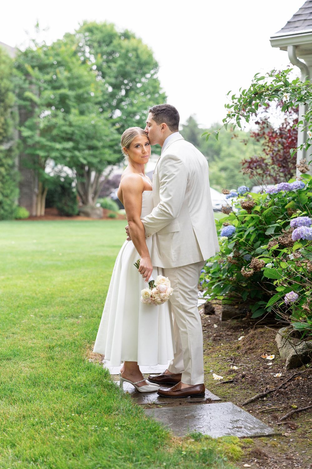 A couple shares a romantic kiss in a garden setting with blooming hydrangeas and lush greenery on a rainy summer day.