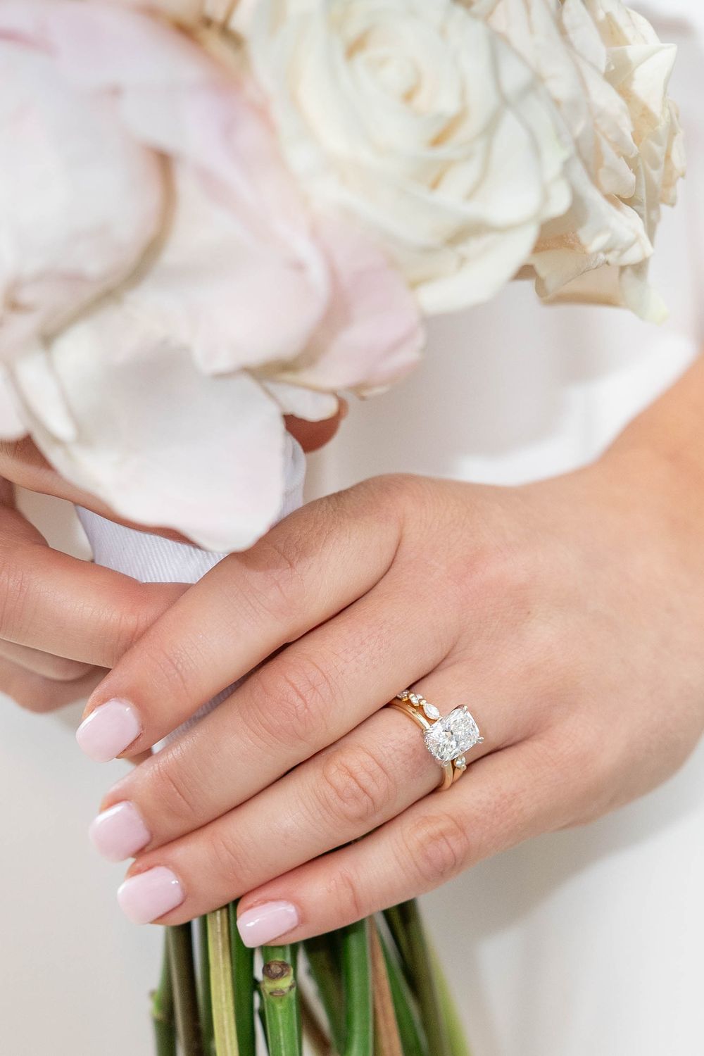 Close-up of a hand wearing an elegant solitaire diamond engagement ring while holding white roses.