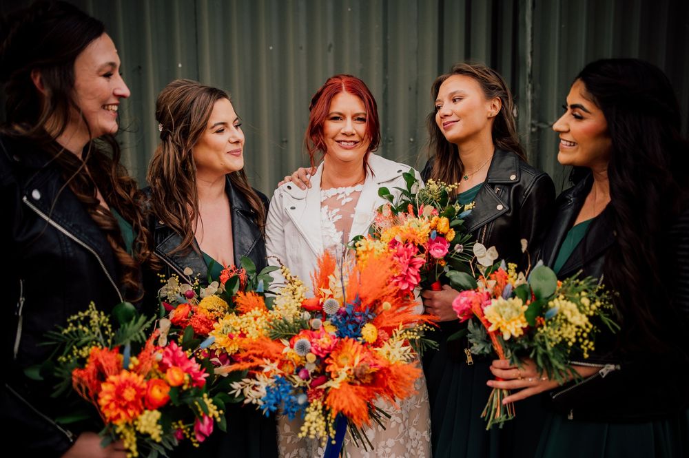 A group of women in colorful dresses laugh and smile while holding vibrant orange and yellow floral bouquets. Worcestershire Wedding photogr