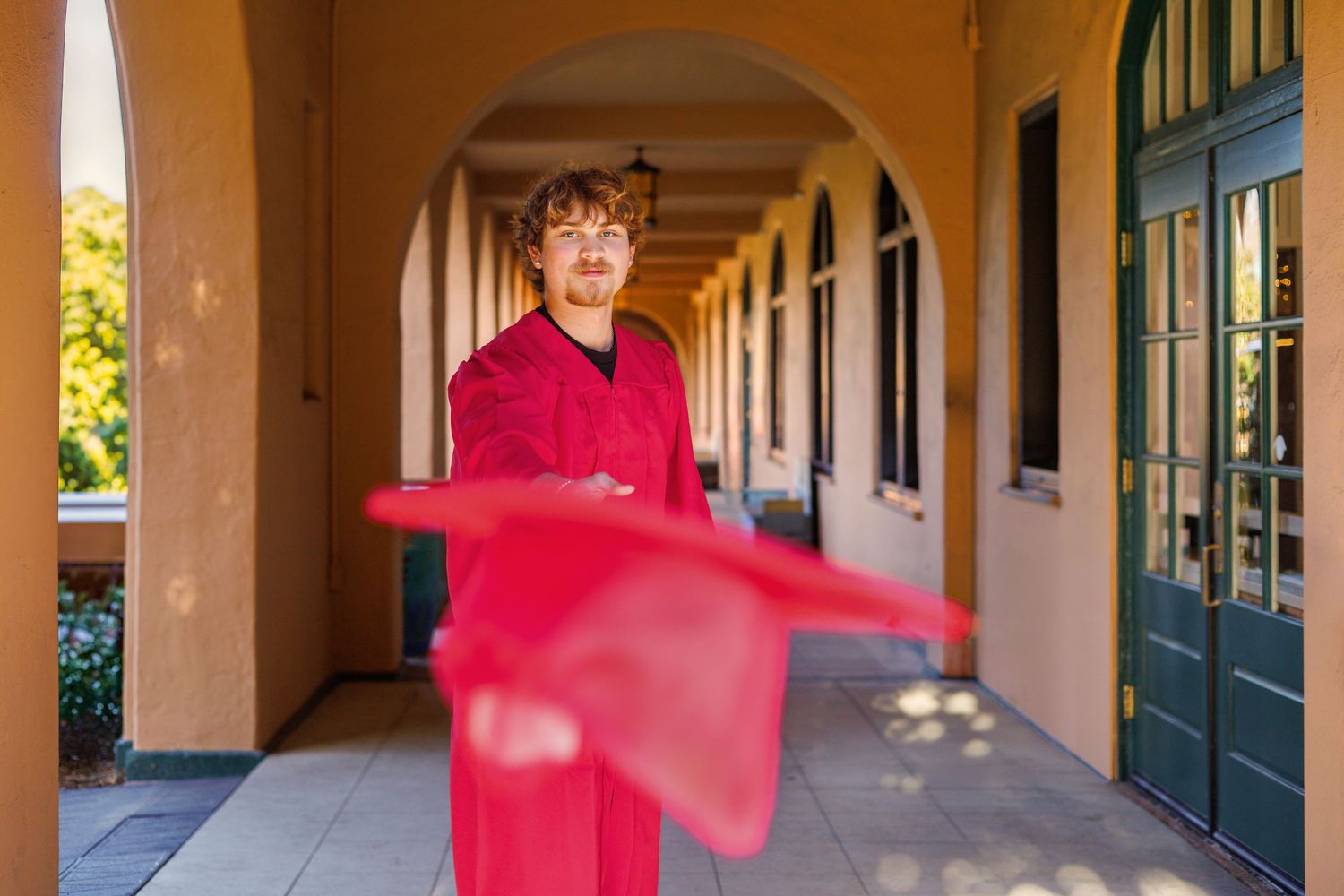 A person in a bright red graduation gown stands smiling in a covered outdoor corridor with arched columns.