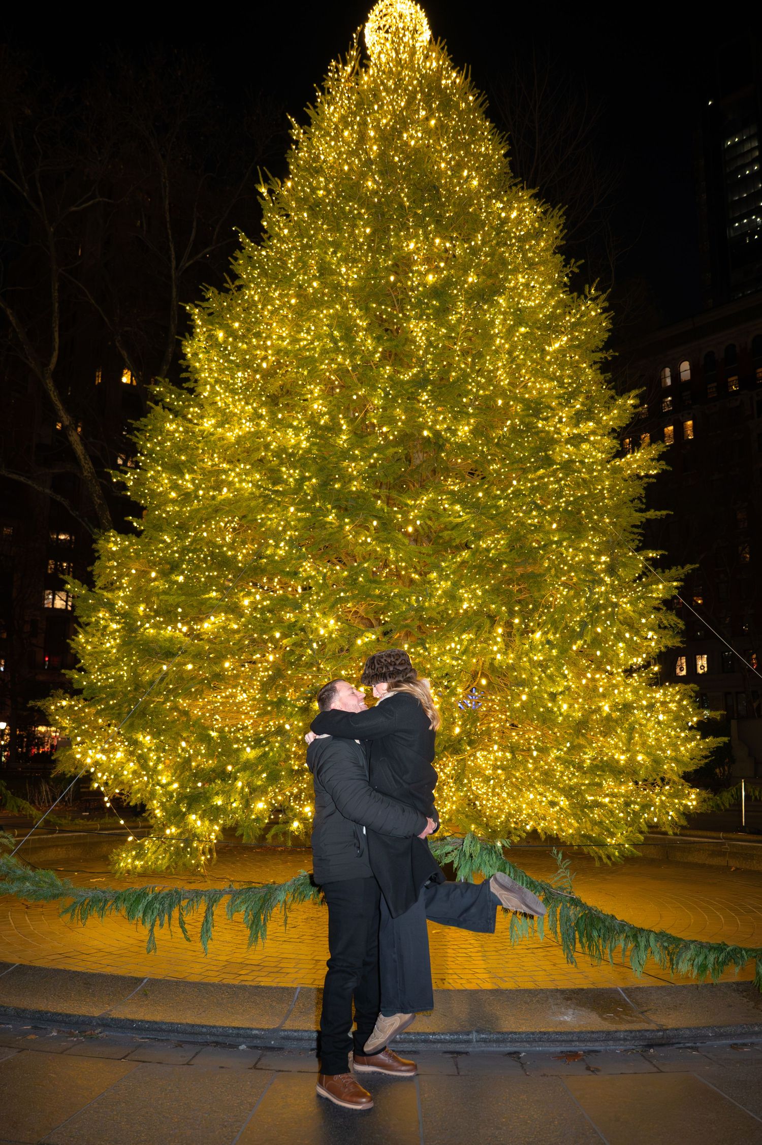 A proposal scene unfolds in front of a beautifully lit Christmas tree at night in Madison Square Park in NYC.