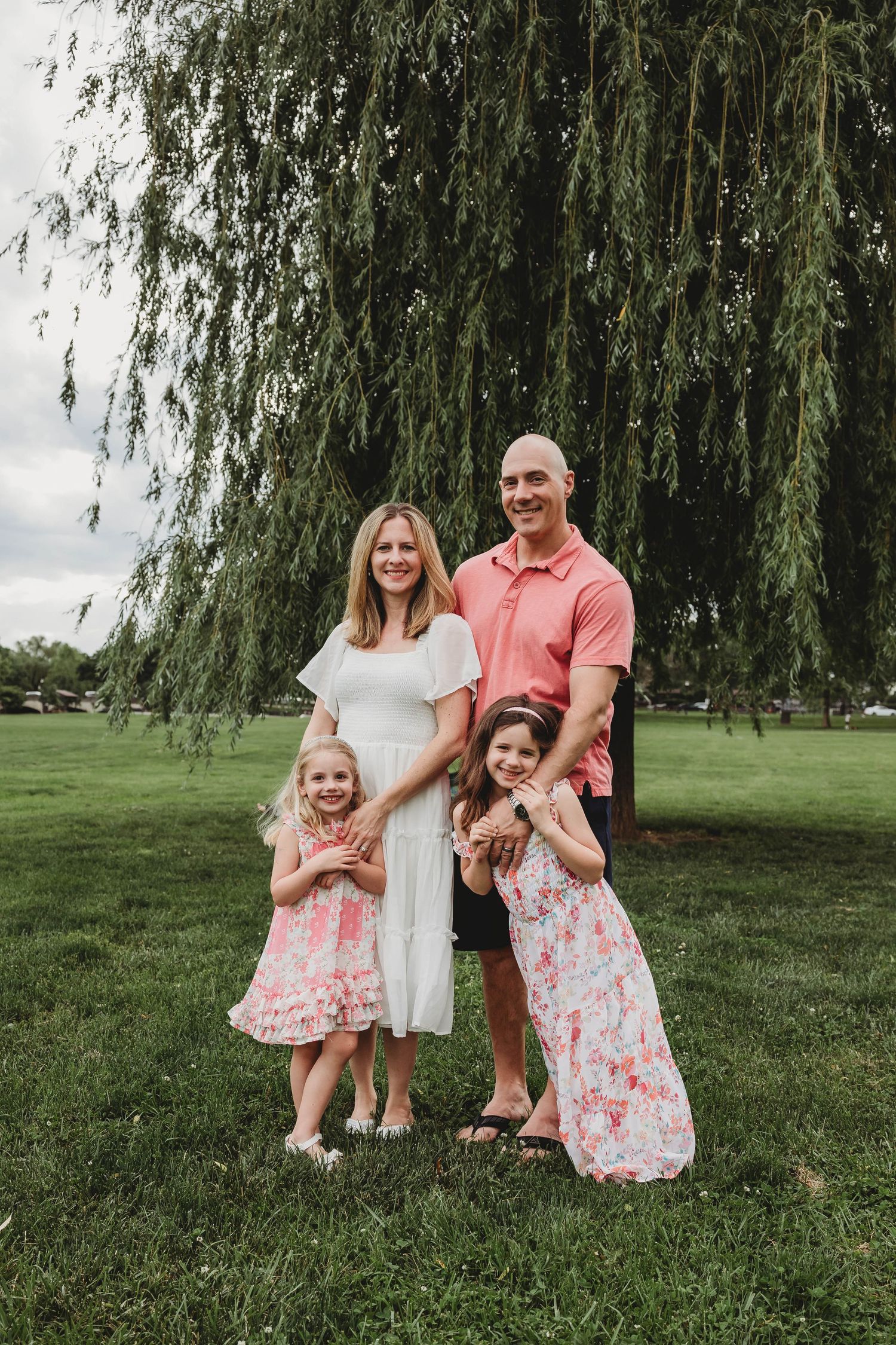 A family of four poses under a weeping willow tree in coordinating pink and white outfits.