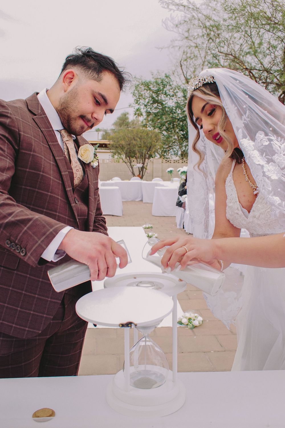 A couple in wedding attire performs a unity ceremony pouring sand together into a clear vessel outdoors.