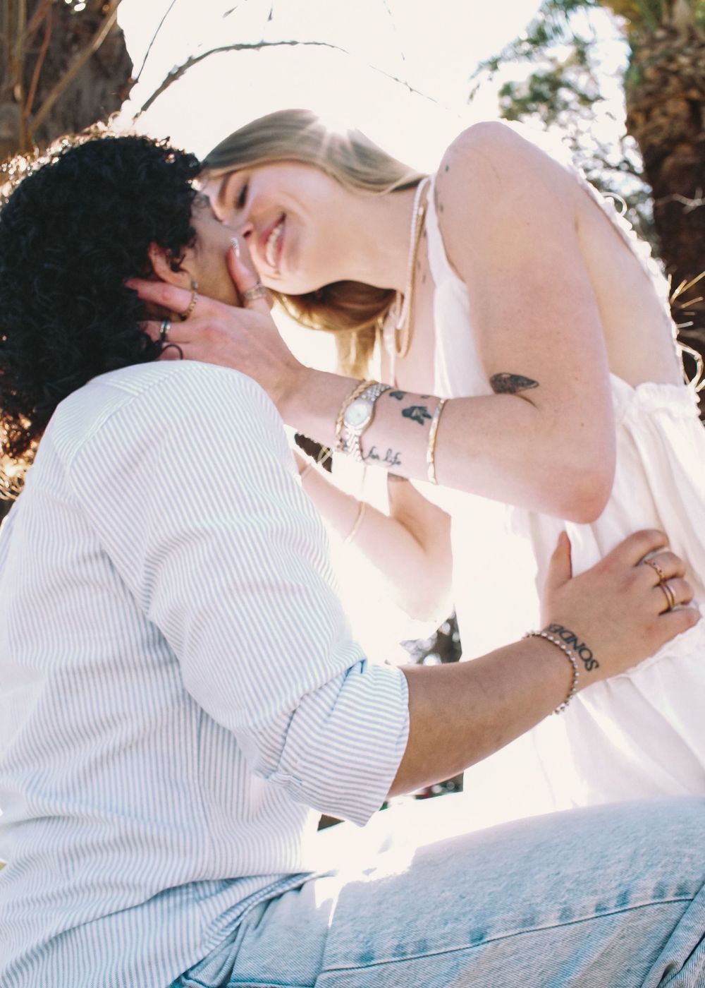 A romantic couple in white clothing shares a tender kiss while sitting on a wooden surface in sunlit outdoor setting.