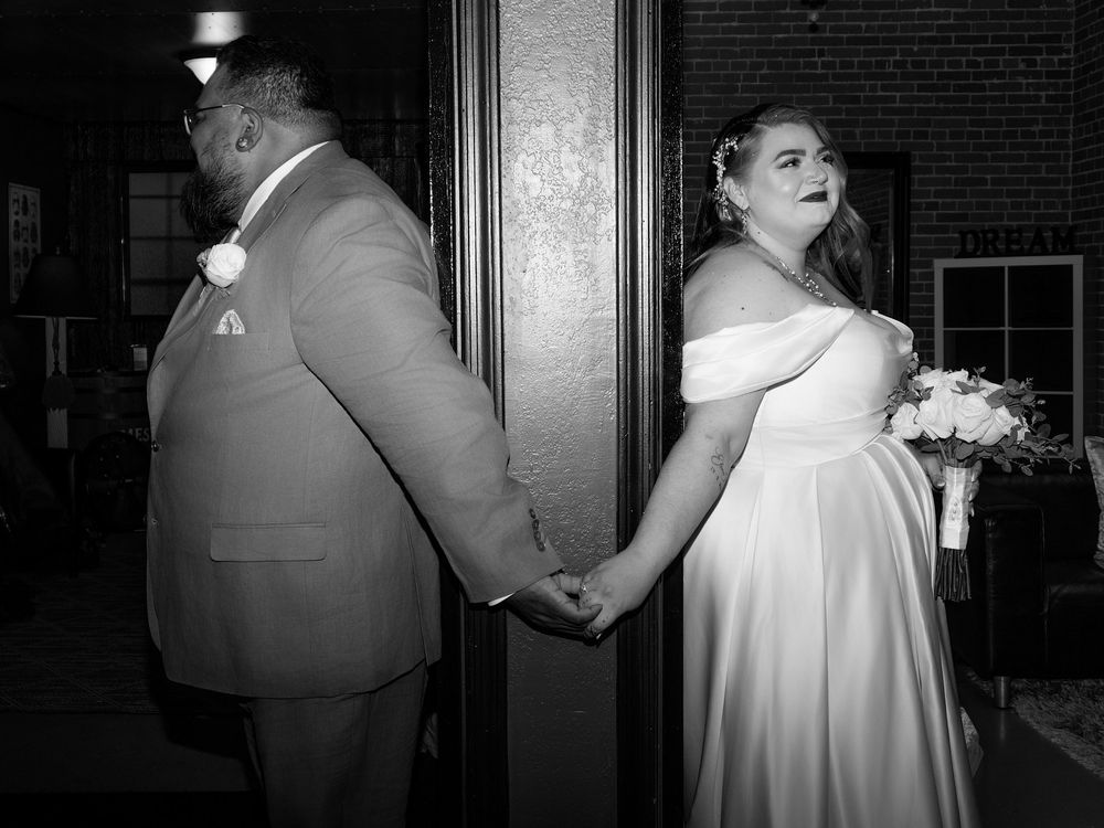 Black and white wedding photo of bride and groom holding hands behind a door, bride in off-shoulder gown holding bouquet.