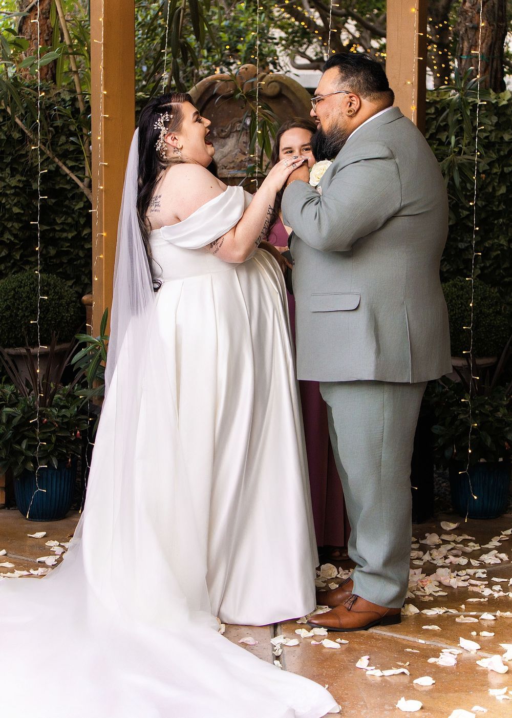 Bride and groom exchange vows at outdoor wedding ceremony under floral arch with rose petals on ground.