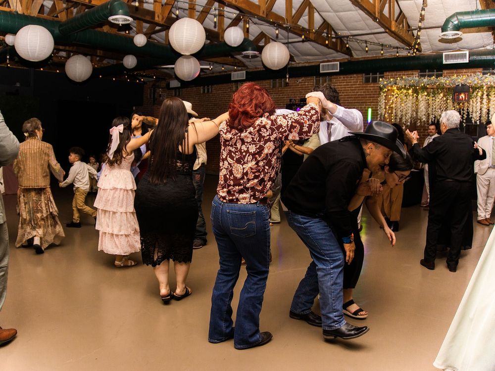 Groups of people dancing together in a rustic decorated hall with white paper lantern lights hanging from the ceiling.