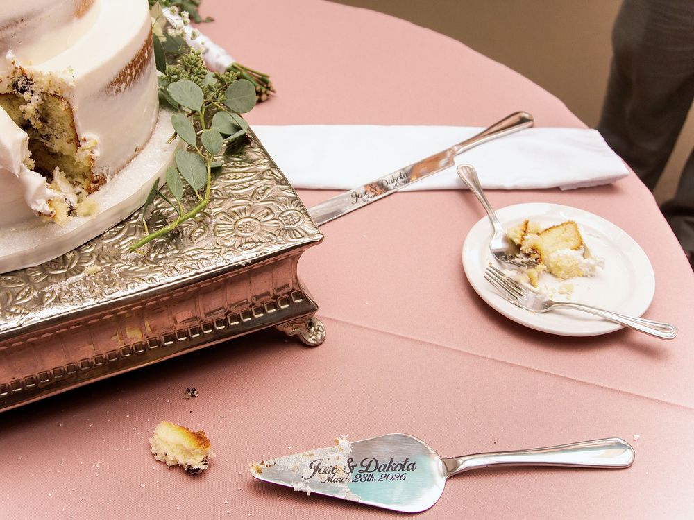 Wedding cake cutting setup with personalized silver server set on a pink tablecloth beside a partially sliced white cake.