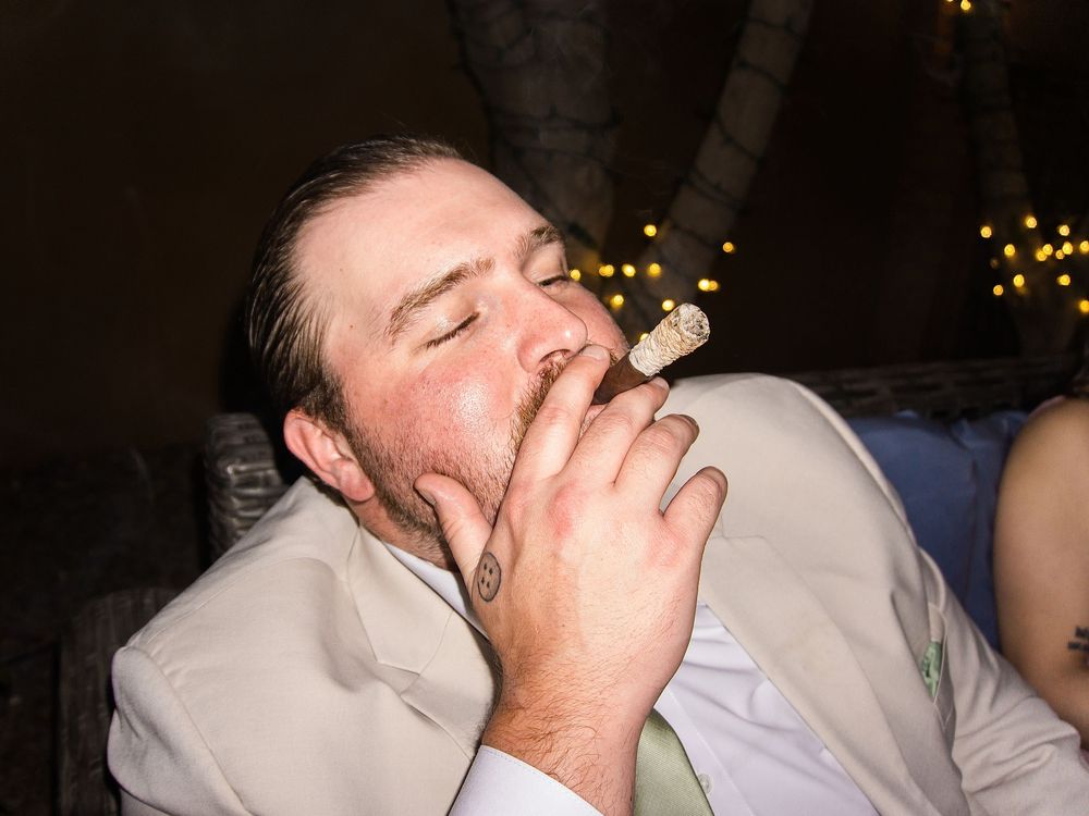 Man in white suit jacket smoking a cigar at an outdoor evening event with bokeh lights in background.