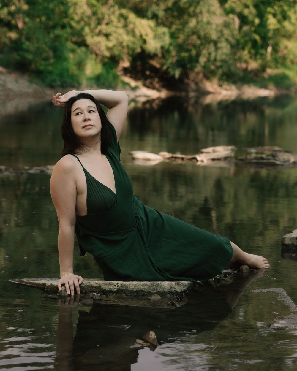 Woman wearing a dark green dress while lying in a river against a rock, looking off to the side