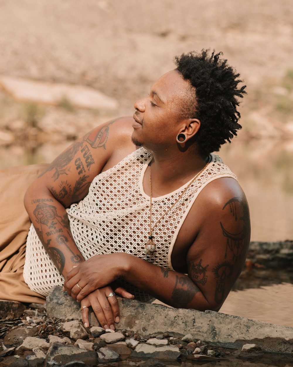 Man wearing a sheer white tank top and a macrame crystal necklace, leaning on the shore of a rocky river with his eyes closed