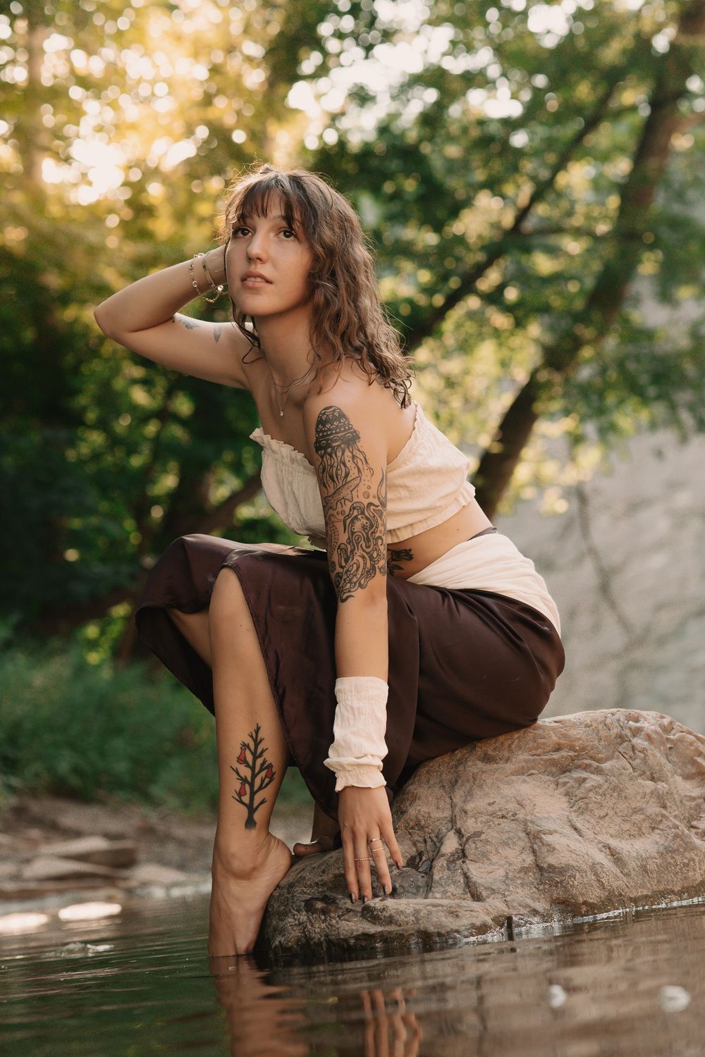 Women wearing a white top and brown skirt while sitting on a rock in a river, holding one hand to the water, and the other hand in her hair