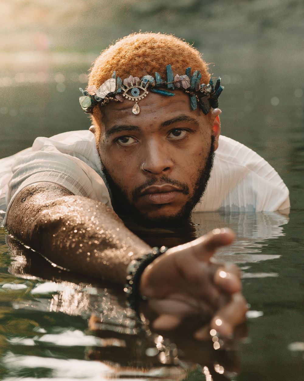 Man wearing a crystal crown swimming in a sun-lit river looking away from the camera