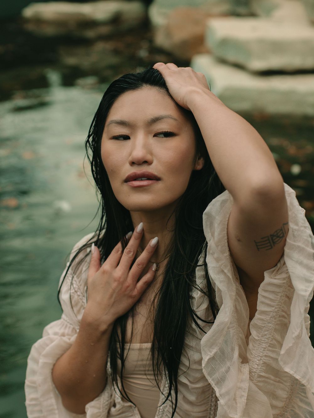 Woman wearing a white, flowy ruffled dress while sitting in a river, holding one hand to her collarbone & the other hand on her head