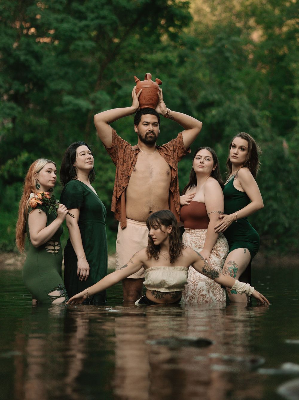 6 people standing and sitting in a river wearing earth toned clothing, playing with the water, holding flowers and a clay pot