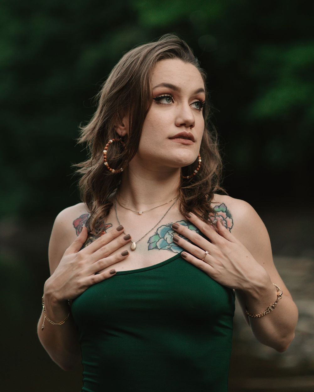 Woman on a riverbank wearing a forest green dress gazing off to the side while holding her hands to her chest