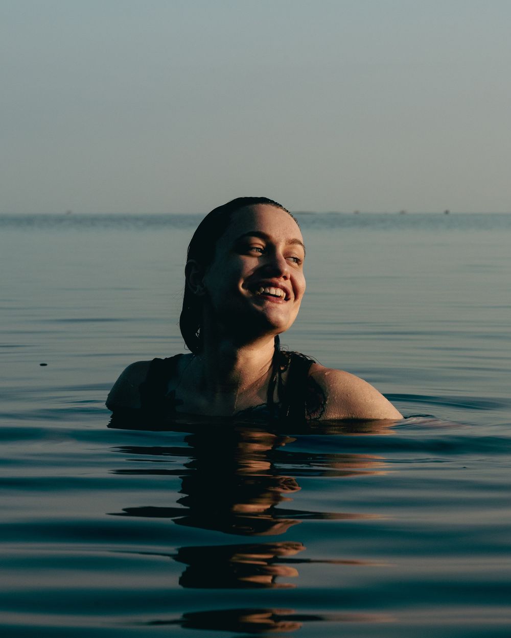 Woman swimming in a calm lake at sunrise surrounded by blue water and the sky, laughing and looking away from the camera