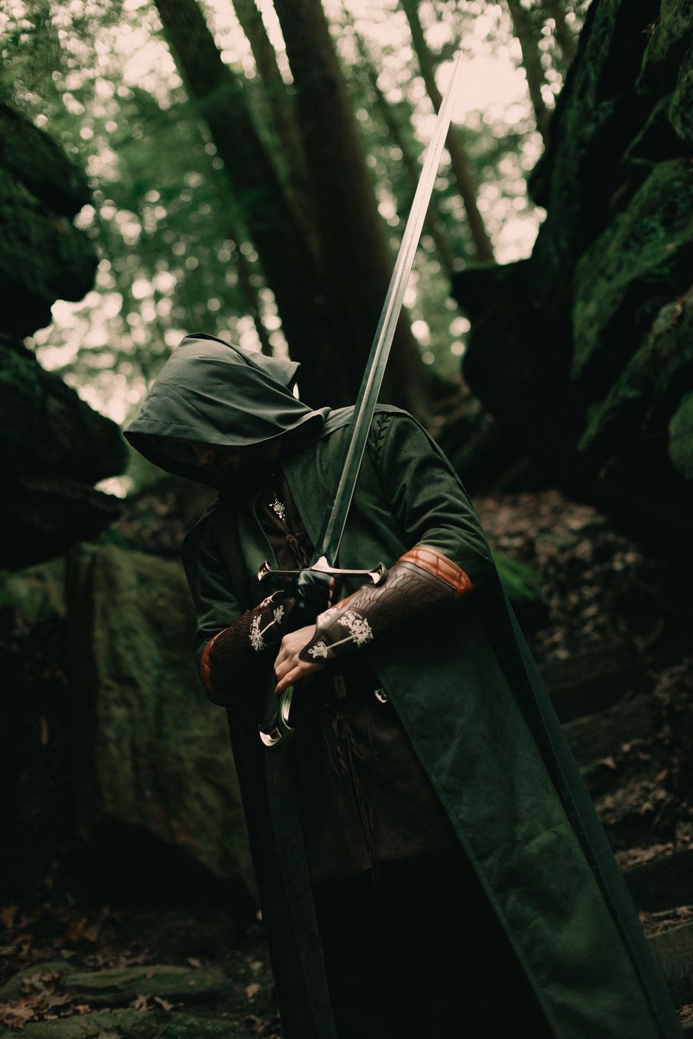Man in a dark mossy forest wearing a cloak and warrior attire holding a sword over his shoulder looking towards the ground